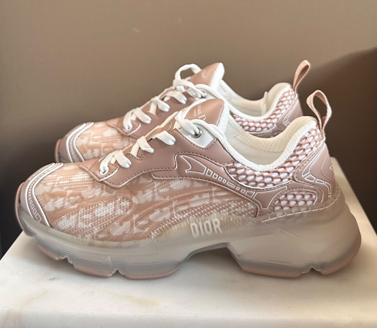 A pair of Dior sneakers in a dusty rose and beige monogram pattern with white laces and a chunky translucent sole, displayed on a light marble surface.