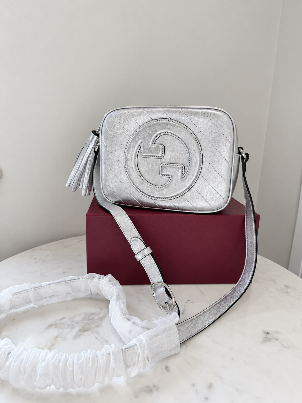    Silver Gucci Blondie bag on its burgundy box with protective wrapping around the strap.