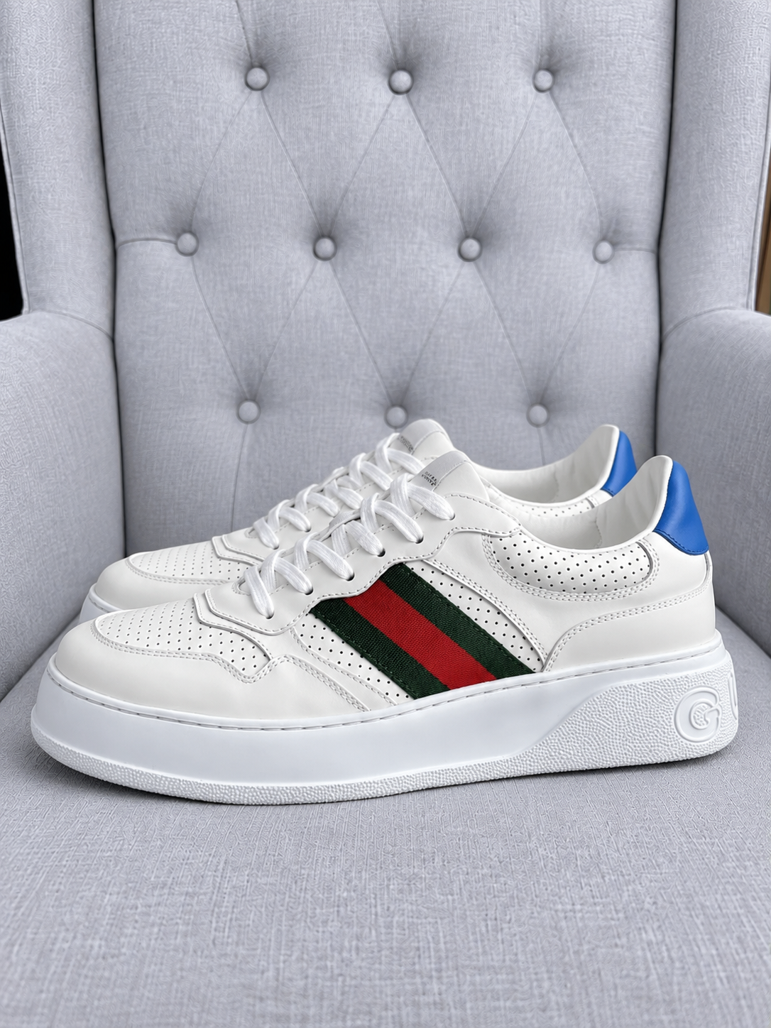 A pair of white Gucci low-top sneakers with the iconic green and red web stripe on the side, a blue heel tab, and a chunky white sole, displayed on a gray tufted armchair.