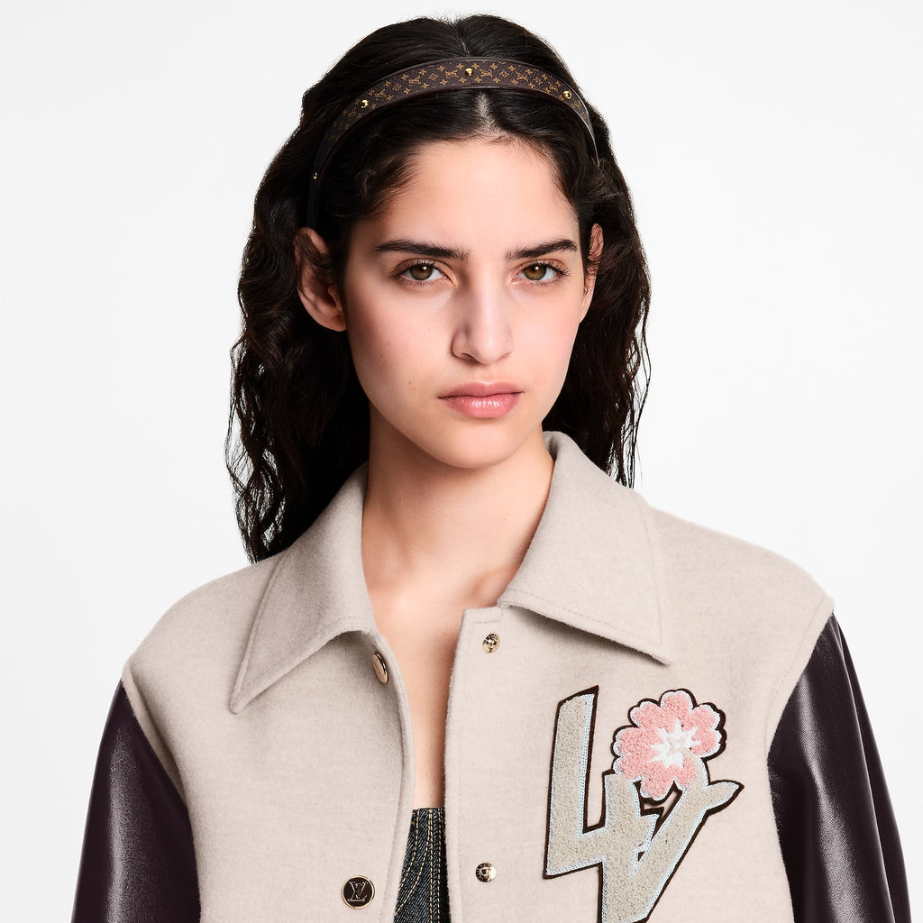 A close-up of a model wearing a beige and black varsity-style jacket with a large LV monogram and pink flower patch on the chest, paired with a black patterned headband.