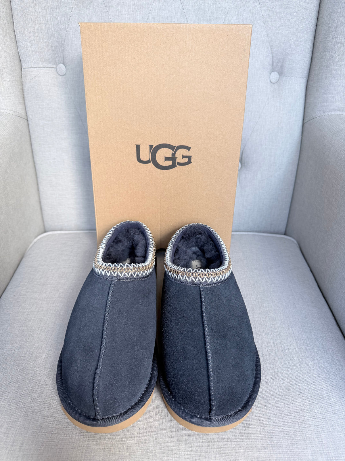 A pair of dark navy blue UGG slip-on slippers with a silver patterned trim and black UGG logo on a brown paper box, placed on a light gray tufted sofa.