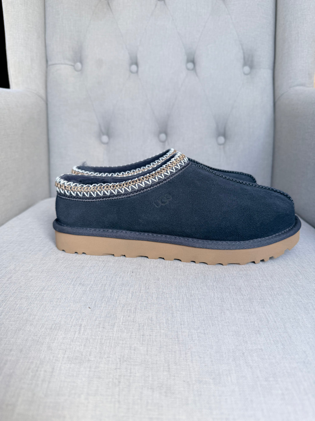 A pair of navy blue suede UGG slip-on clogs with a decorative white and gold chevron trim around the collar, resting on a light gray tufted armchair.